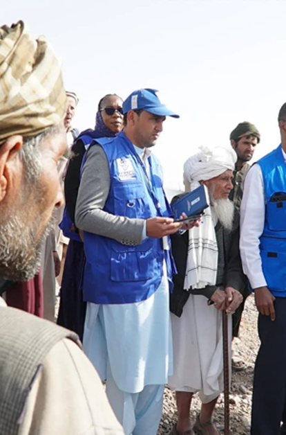 Photo: Group of people outdoors, some in blue vests, listening to a speaker on a dusty field.